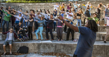 ‘The Complete Freedom of Truth’ project in Bryanston School in Dorset, UK. A mixed group of people dance in an outdoors amphitheatric setting. Their hands are raised toward their left as they lean their bodies to the right.